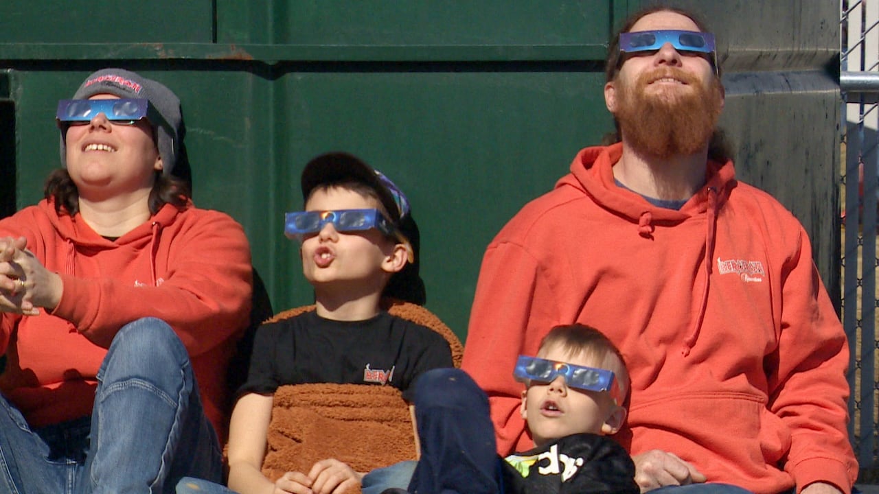 A woman, two kids and a man sat against a wall. All of them wear eclipse glasses.