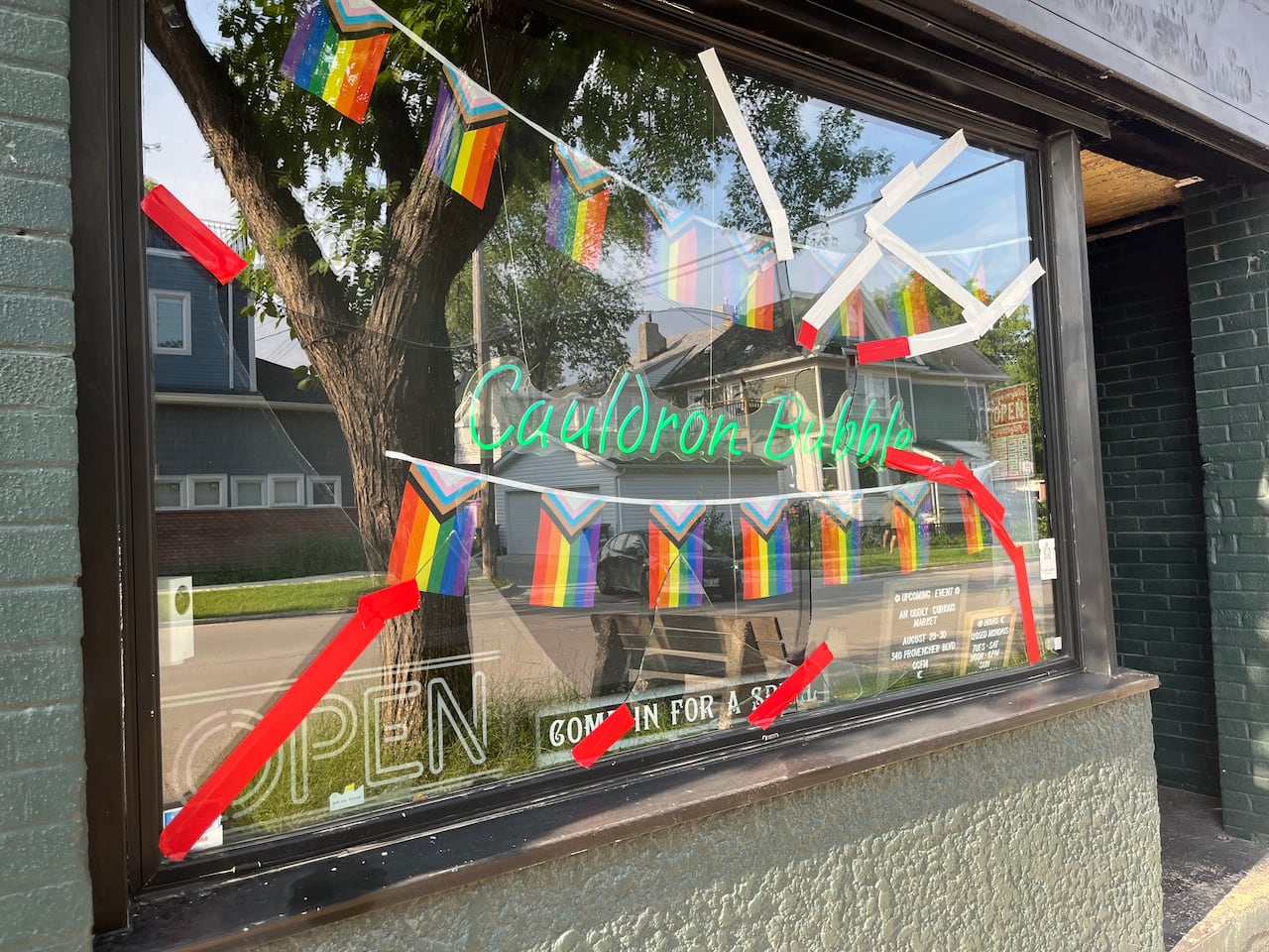 A glass storefront with a neon sign reading "Cauldron Bubble" and rows of Pride flags across it is patched up with red and white tape.