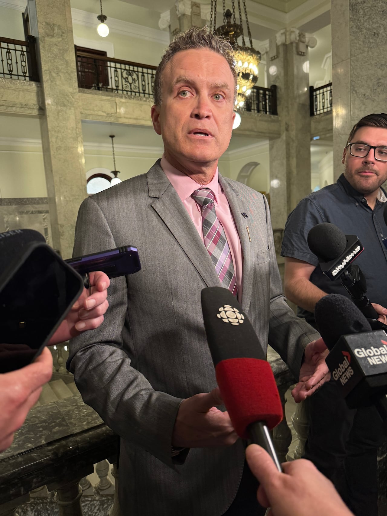 A man wears a grey suit and is surrounded by reporters holding microphones inside the Alberta legislature.