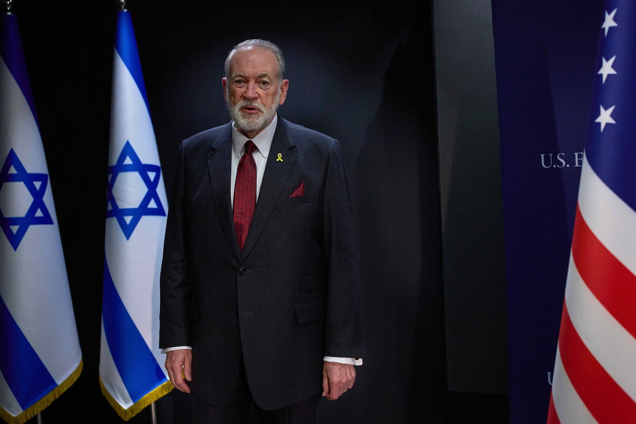 a bearded, balding man in suit and tie is shown standing near American and Israel flags.