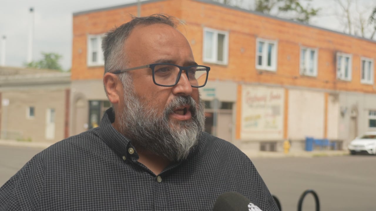 Side shot, close up of Rino speaking into a reporter's microphone with a brick lowrise building in the background.