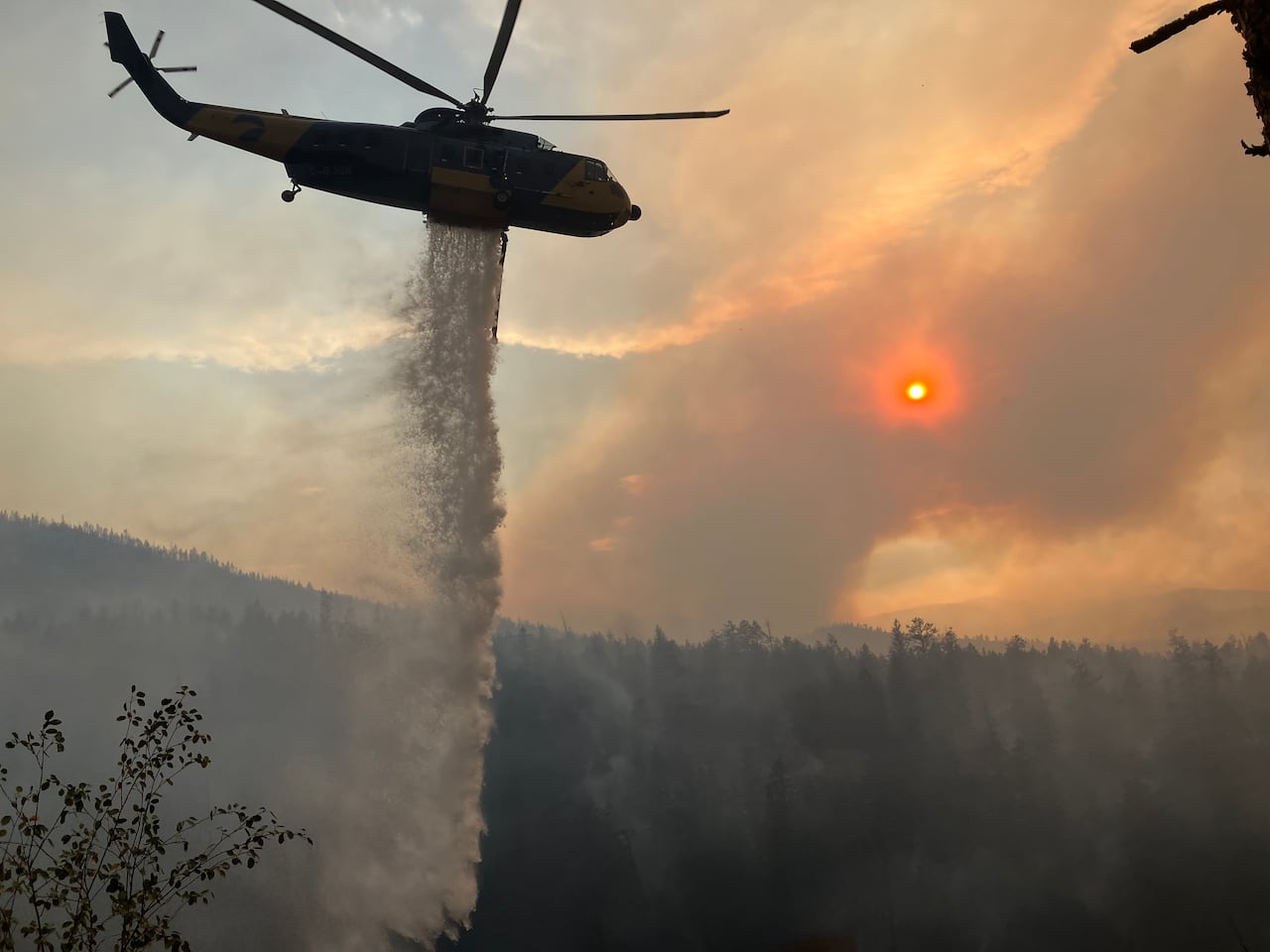 A helicopter is pictured dumping water above a smoky forest with a red sun in the background.