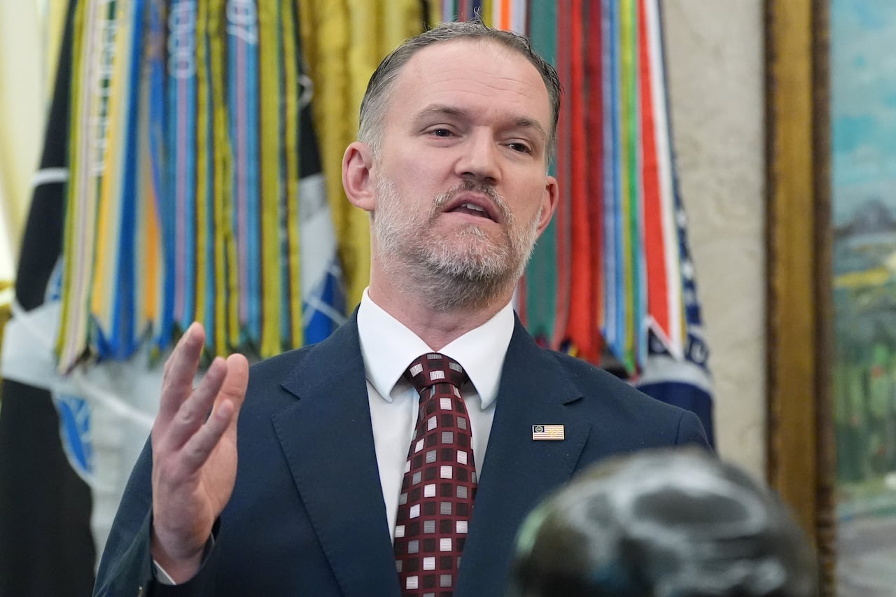 A man in a navy suit with a checkered tie gestures with his right hand while speaking.