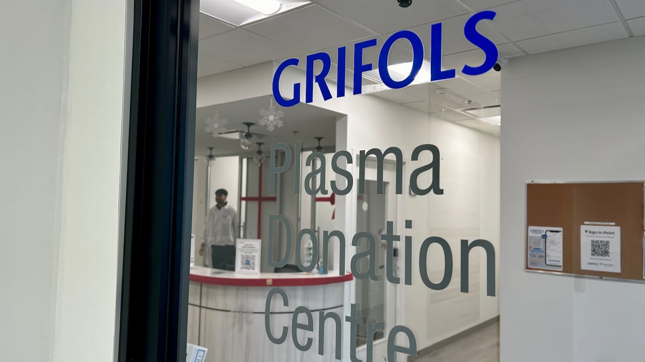 A glass door with the words Grifols Plasma Donation Centre, and a blurry man in the background walking toward a reception desk. 