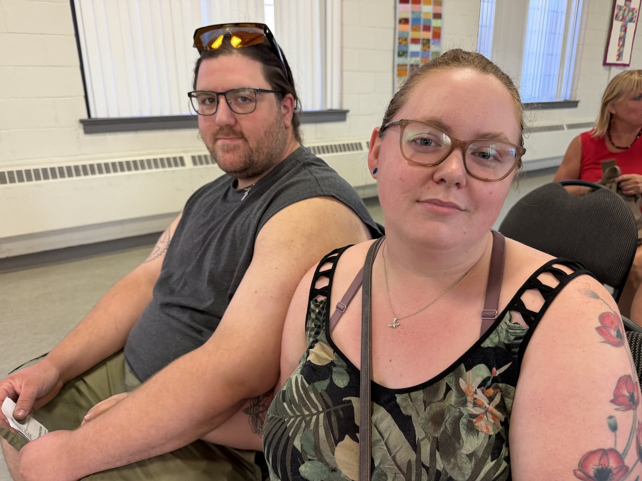 Two people sit next to each other and face the camera. One wears a gray top, the other green floral. Both wear glasses.