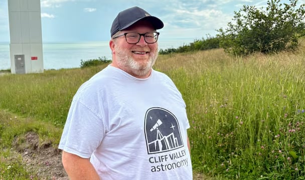 Man, standing and looking at camera, on a sunny day, with a lighthouse behind him.