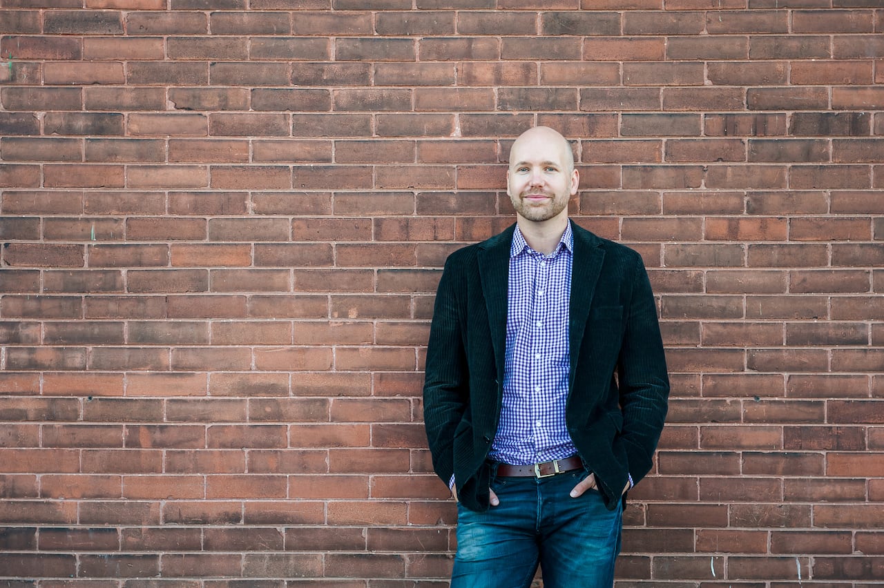 Man wearing blazer with hands in his pockets stands in front of brick wall