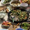 This photo shows a green bean and chicken stir-fry, with sauce and seasonings, in a black pan. It is surrounded by other dishes that include vegetables and rice.
