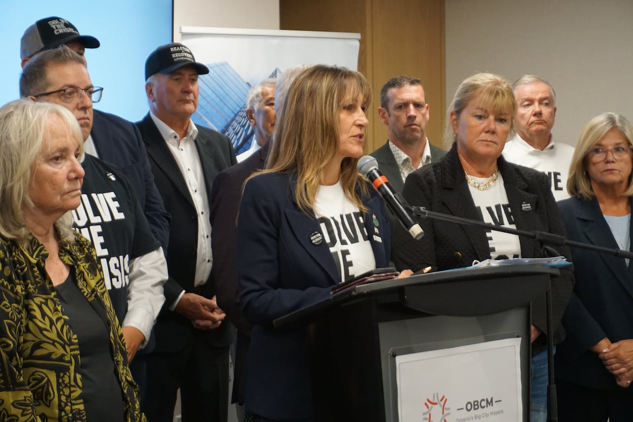 A woman stands at a podium and speaks into a microphone while surrounded by about a dozen other people.