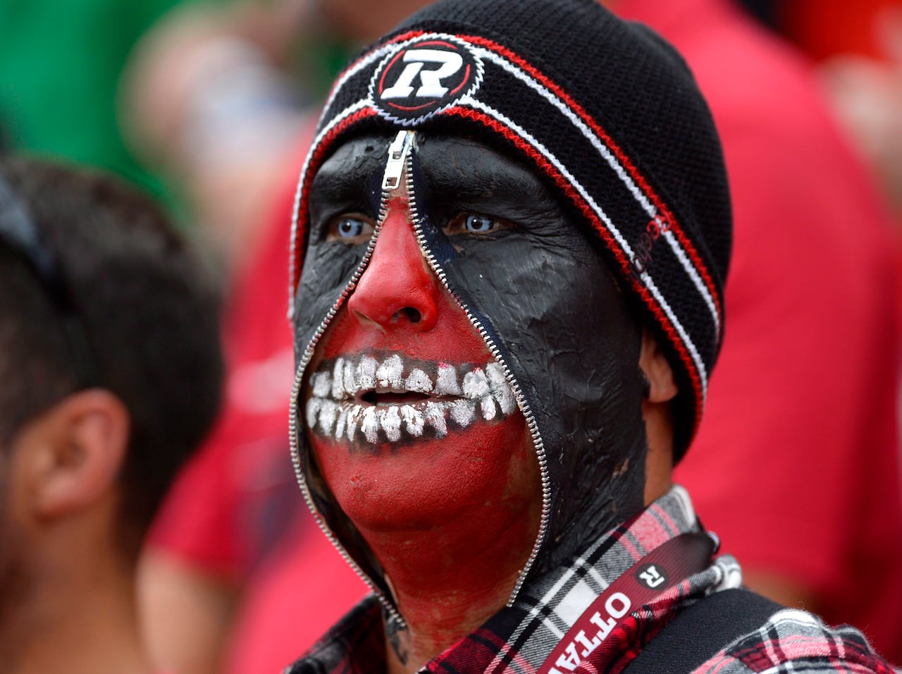 A man whose face is painted red and black with a zipper running up the middle and exaggerated painted teeth.