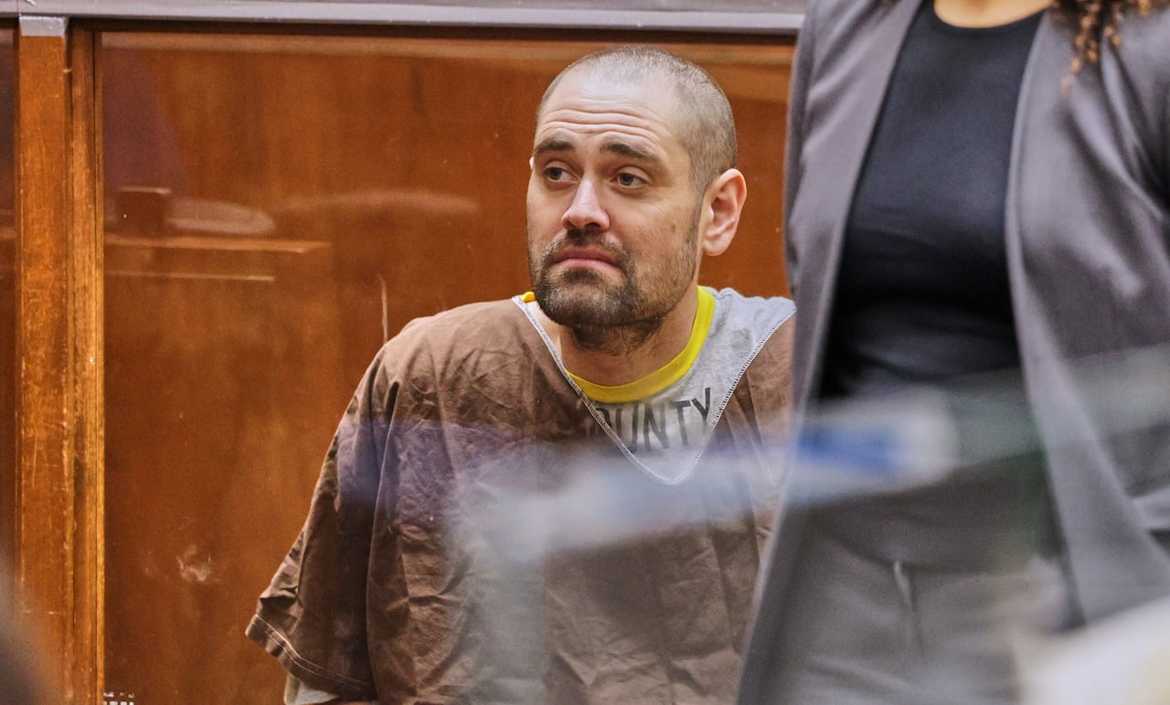 A man sits in a courtroom. A woman is standing in front of him, partially blocking him.
