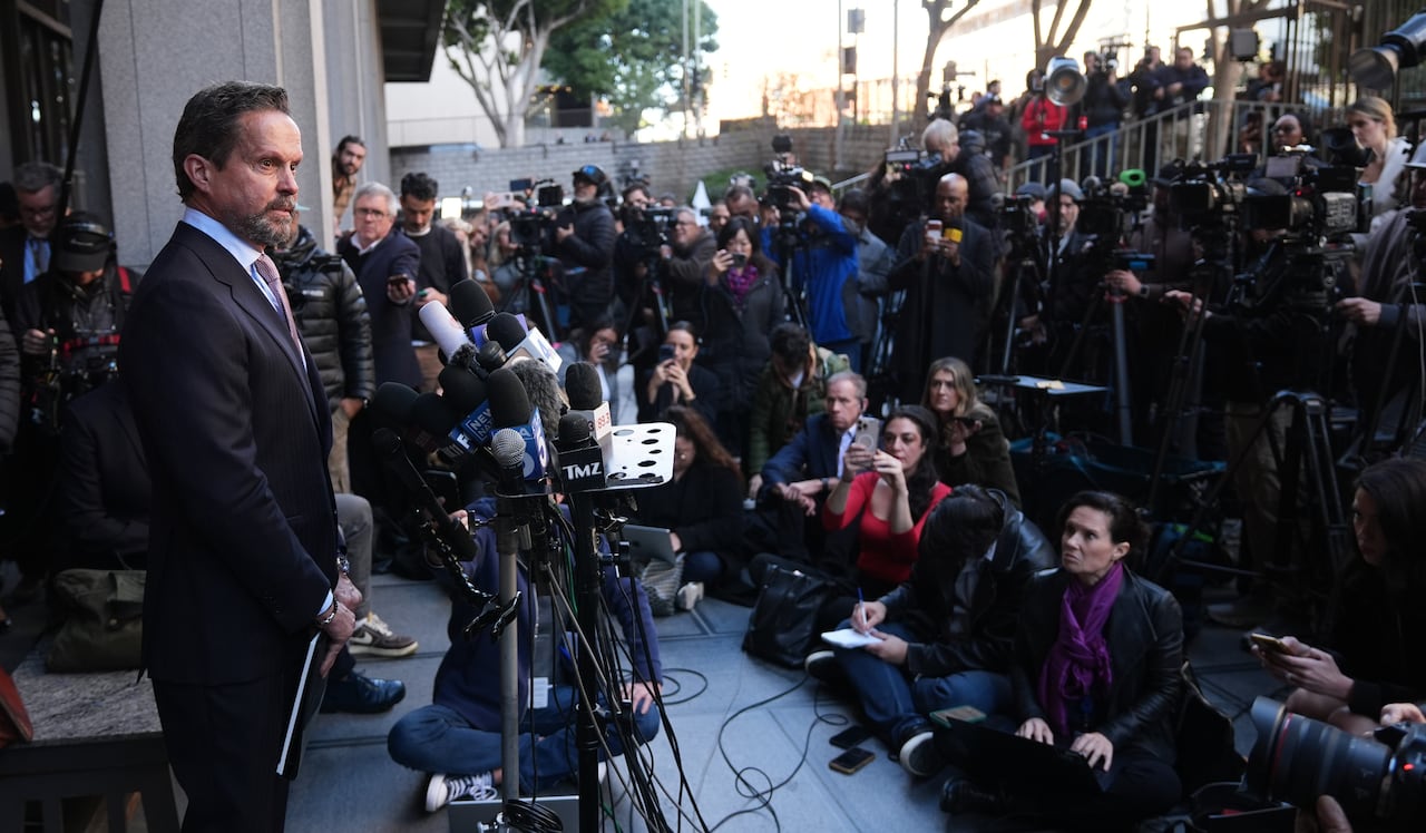 A man in a suit stands outside a building at a microphone, facing a large crowd of reporters, some of whom are sitting on the ground.