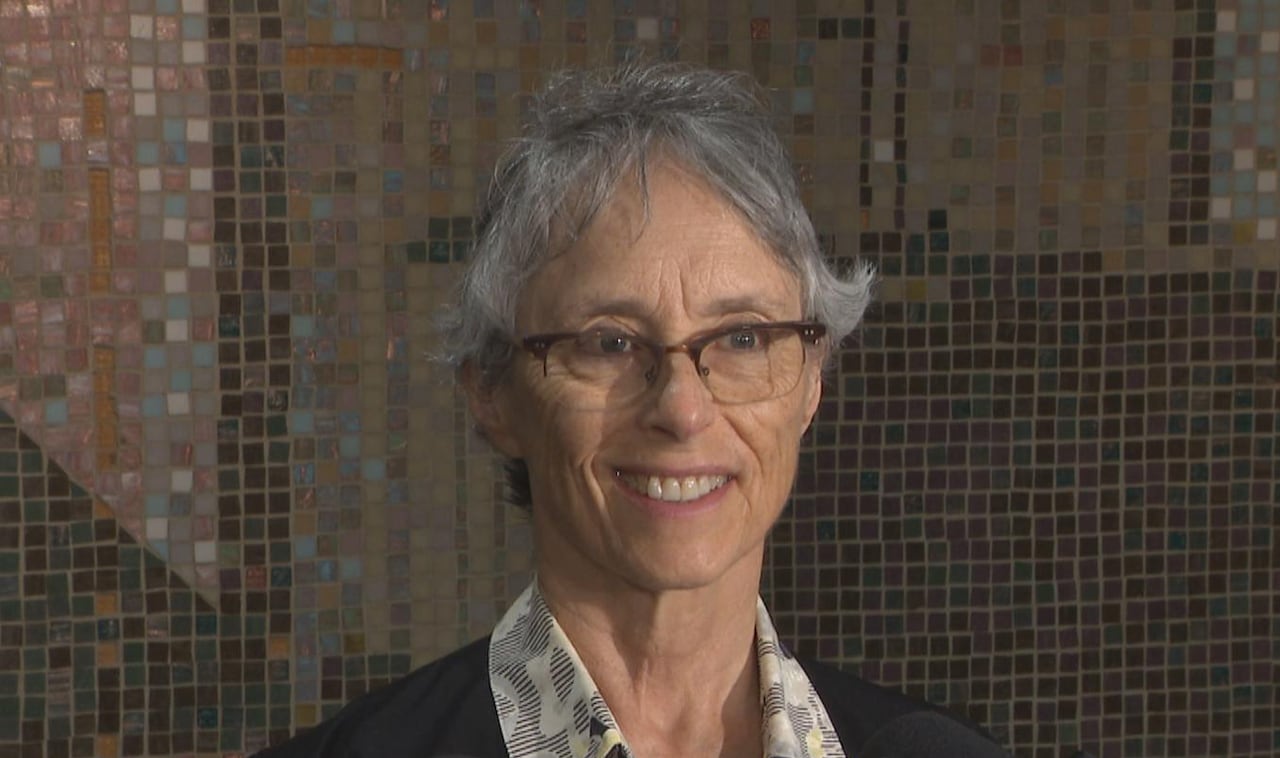 Woman in button-up shirt and black blazer stands in front of microphones, smiling