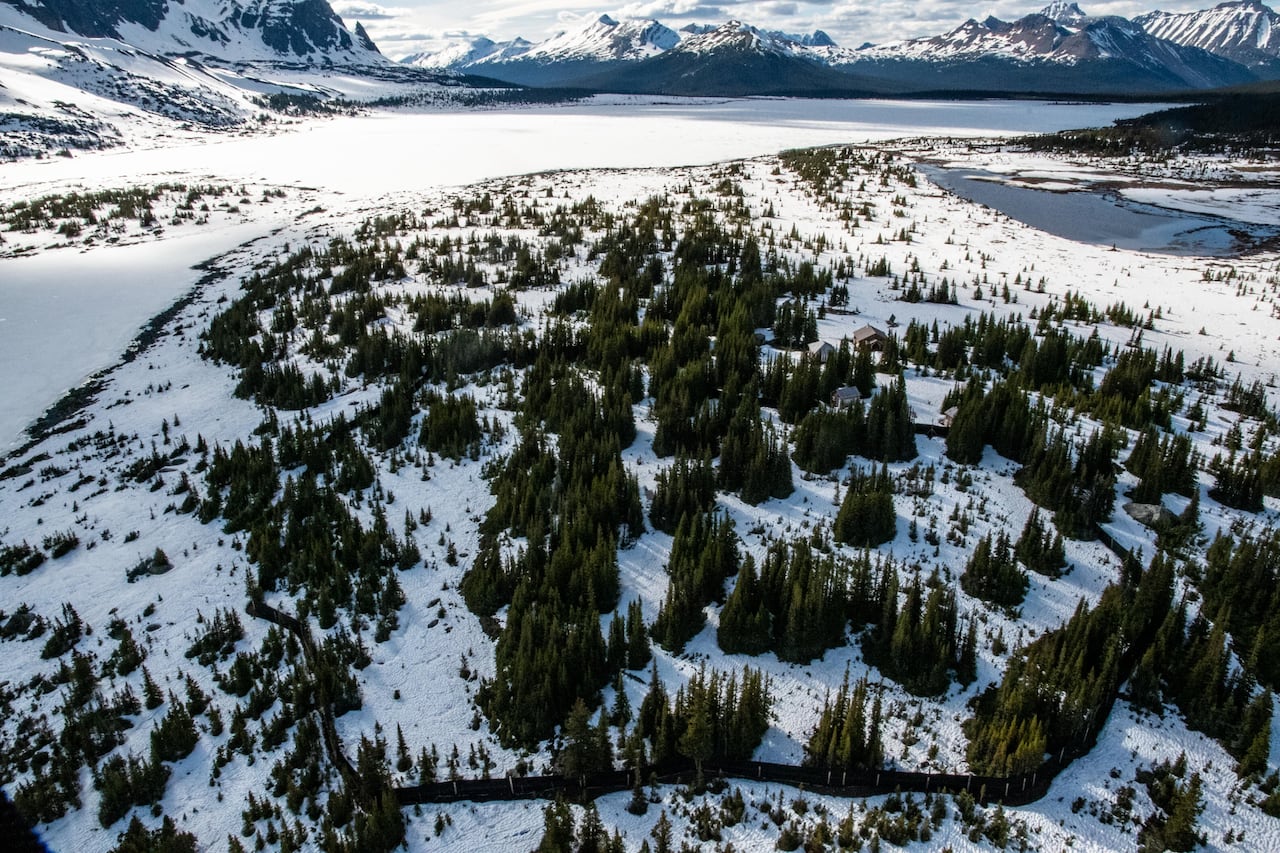 An aerial shot of a frozen lake and mountain landscape. A black fence surrounds a portion of the snowy wooded area.