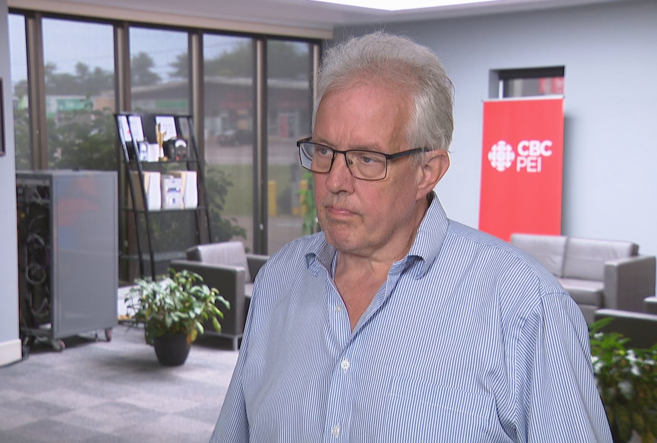 a man wearing glasses and a blue button-up shirt standing indoors near a CBC PEI sign