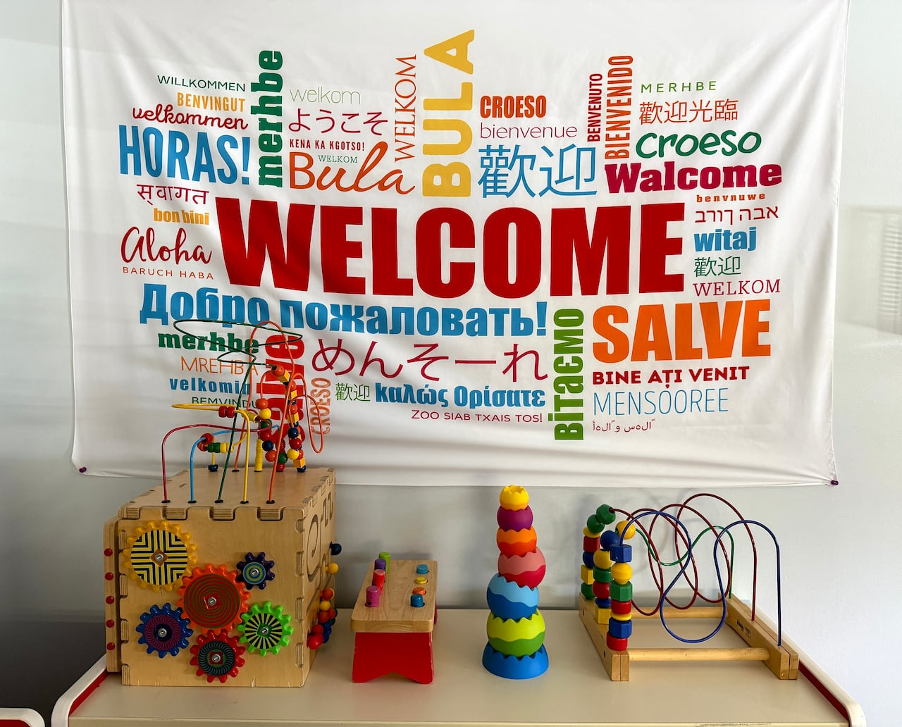 A white banner with multi-coloured text reads "welcome" in English and many other languages. Below, a table contains a number of puzzle toys for small children.