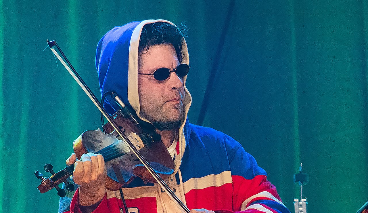 Two people play fiddles onstage in a performance. The man on the left is wearing a hockey jersey and sunglasses, while the man on the right is wearing a suit.