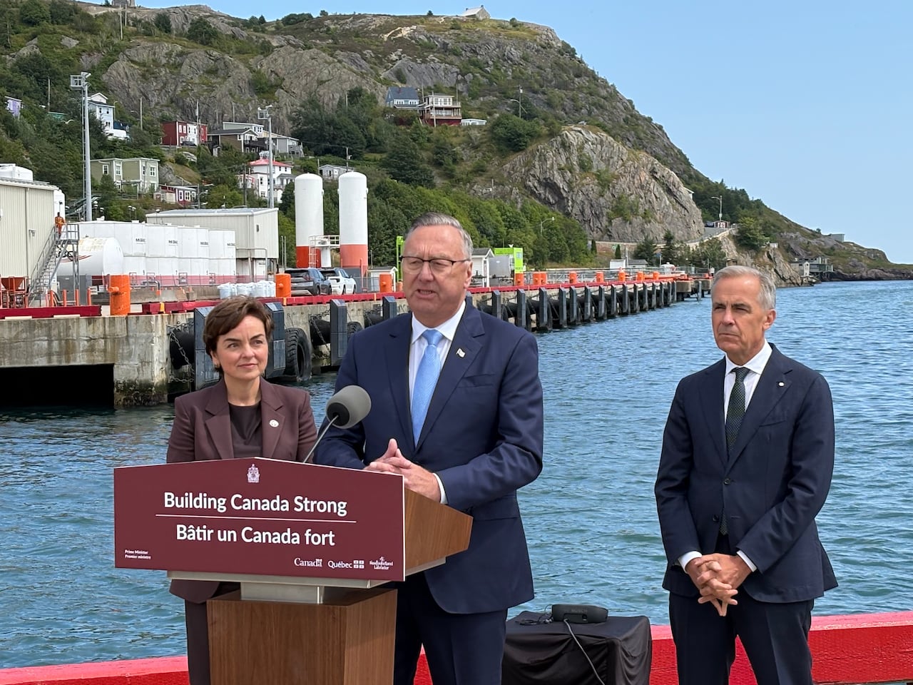 Three people standing, one in front of a podium that says "building Canada strong," against a backdrop of the ocean.