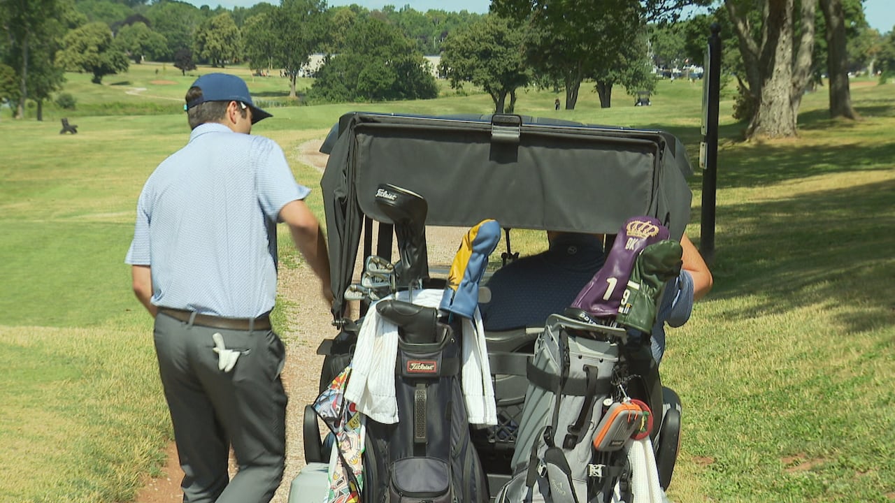 A man climbs into a golf cart on a course. There are two golf bags filled with clubs in the back of the cart.