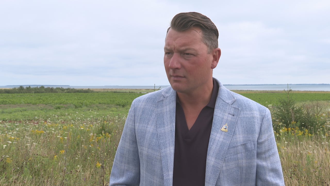 A man in a blue suit stands among greenery with water in the background.