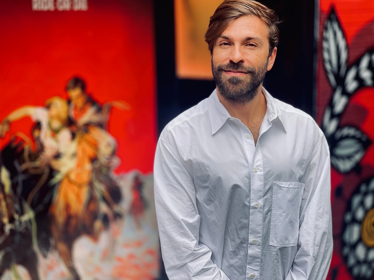 A man with brown hair and a blue shirt stands in front of his art gallery.
