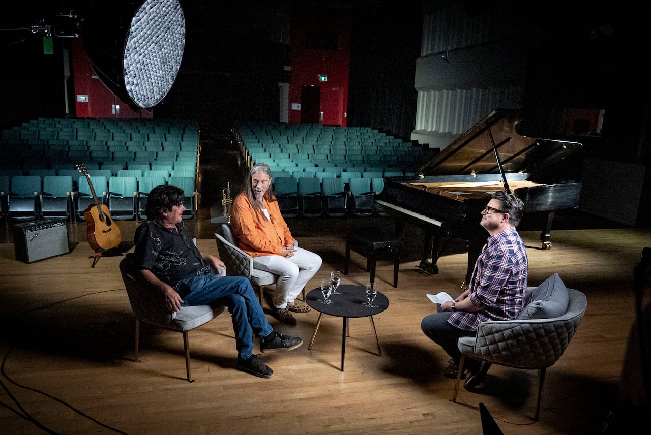 Three men sit in chairs with broadcast equipment at a theatre.