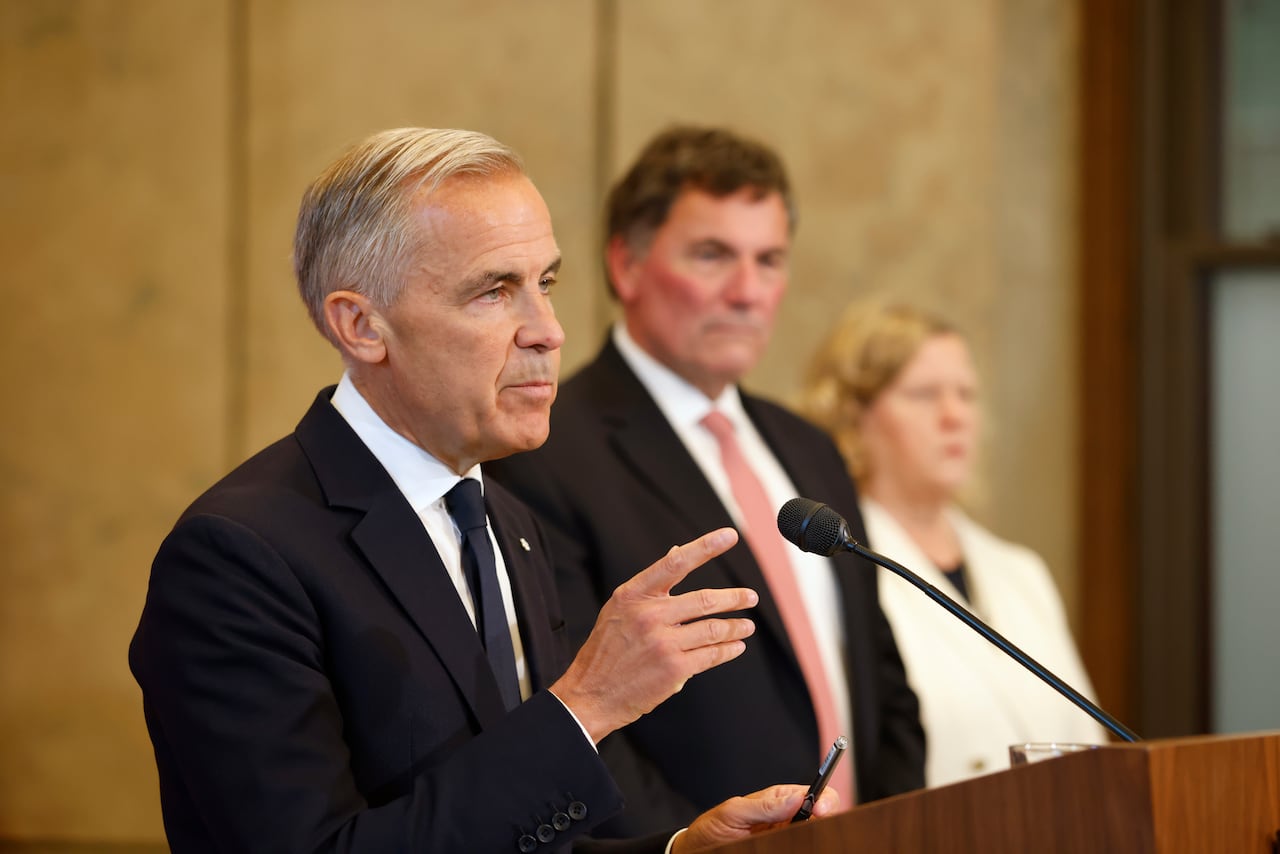 A man in a suit speaks at a podium as another man in a suit and a woman in a white business suit watch.