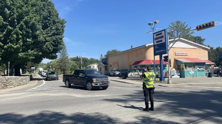 A police officer stands on a road on a sunny day directing traffic in front of a long lineup of cars and trucks.