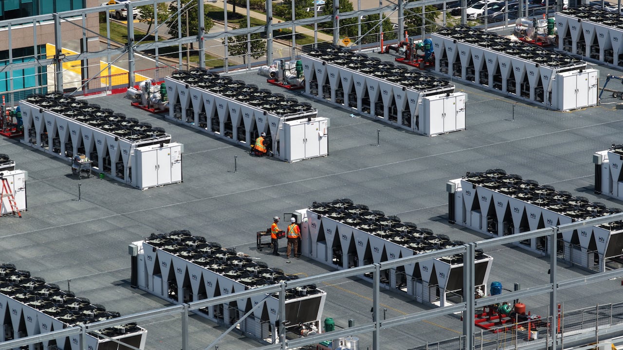 Drone shot of the roof of a data centre, with rows of what appear to be giant fans, a few people in orange construction outfits dotted around.