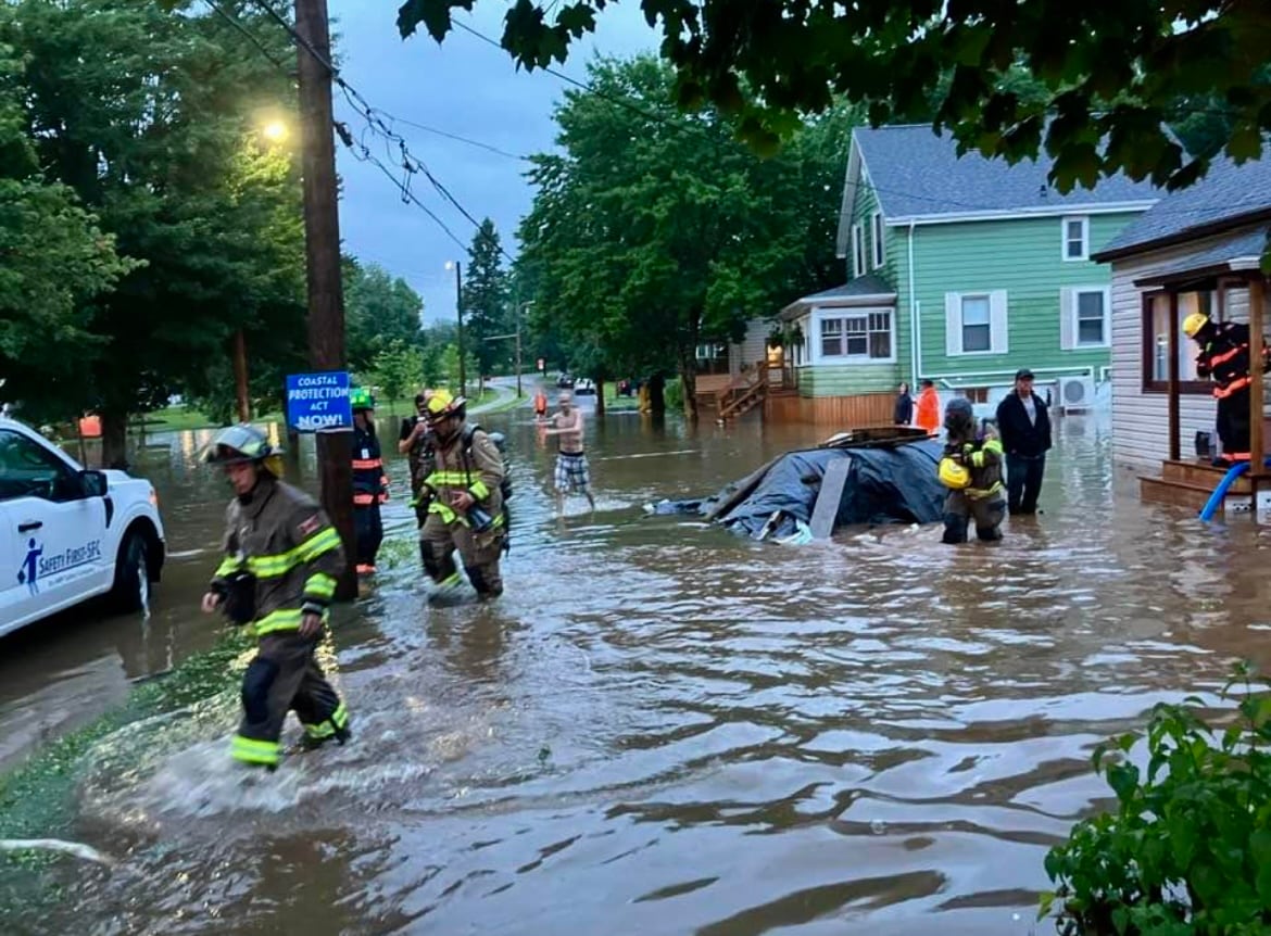 Firefighters are walking through a couple feet of water on a flooded residential street. 
