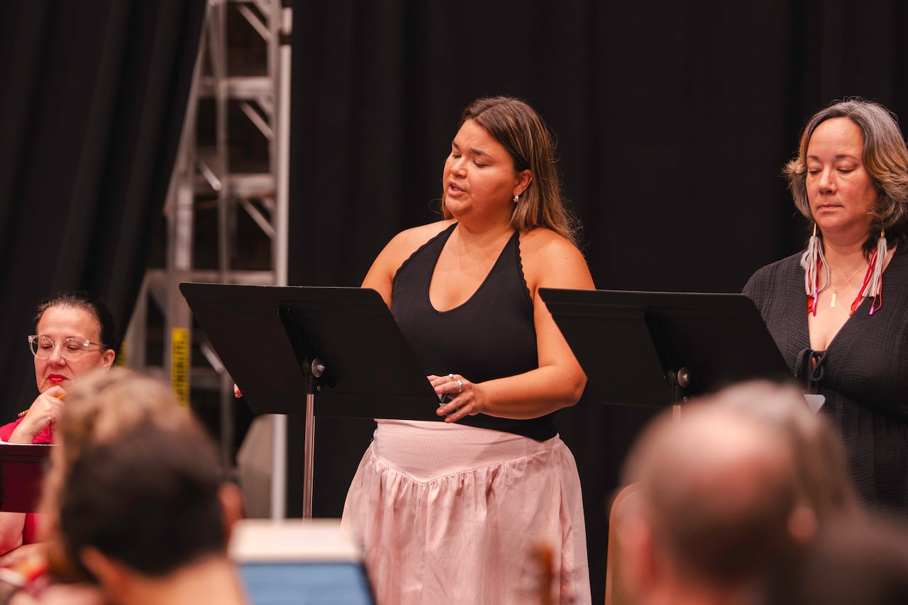 A woman in a pink dress and black halter top sings with a music stand before her. There's another woman standing beside her behind another music stand.