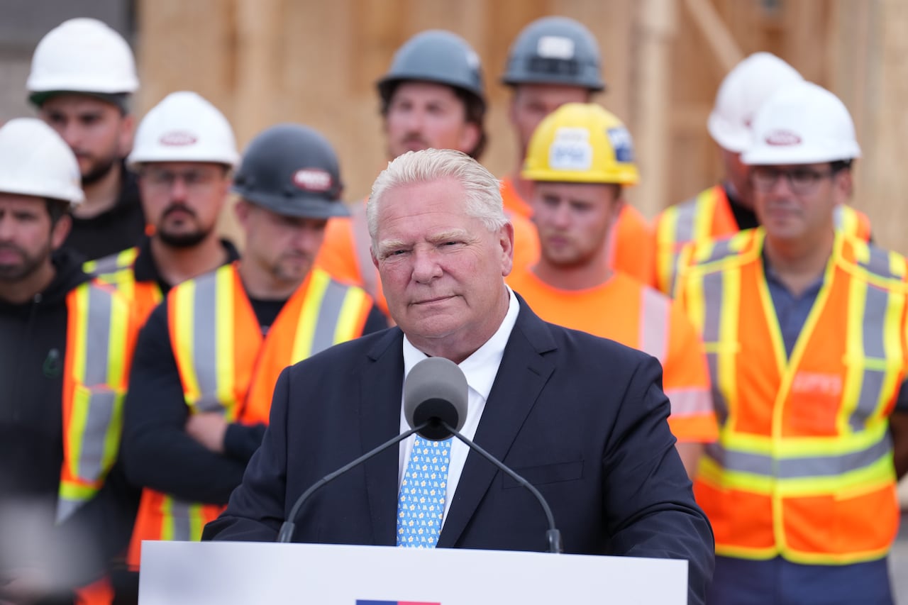 A man stands at a podium with a microphone in front of construction workers wearing bright orange vests, wearing a dark navy suit.