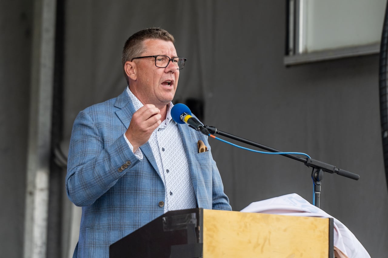 A man in a white and blue shirt stands at a lectern and speaks into a microphone.