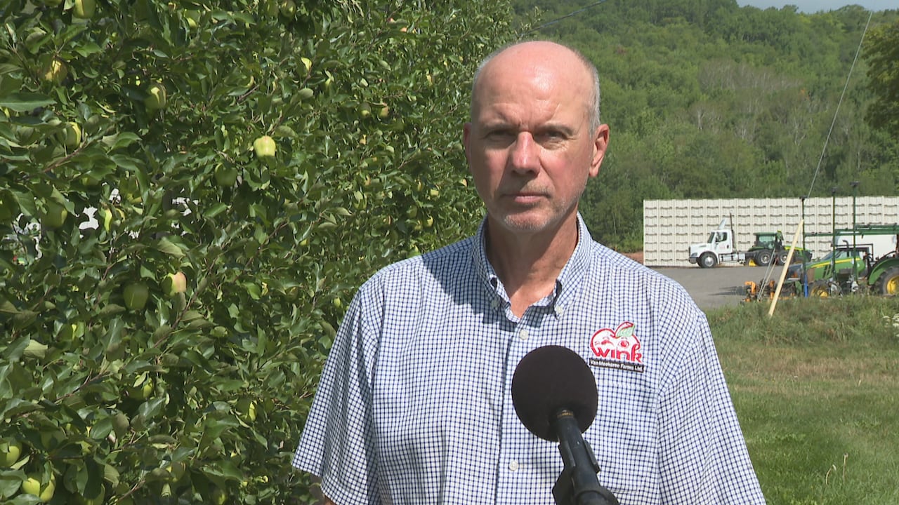 A man standing in front of a microphone. Next to him, a few apples trees. Behind him on the back, a truck,