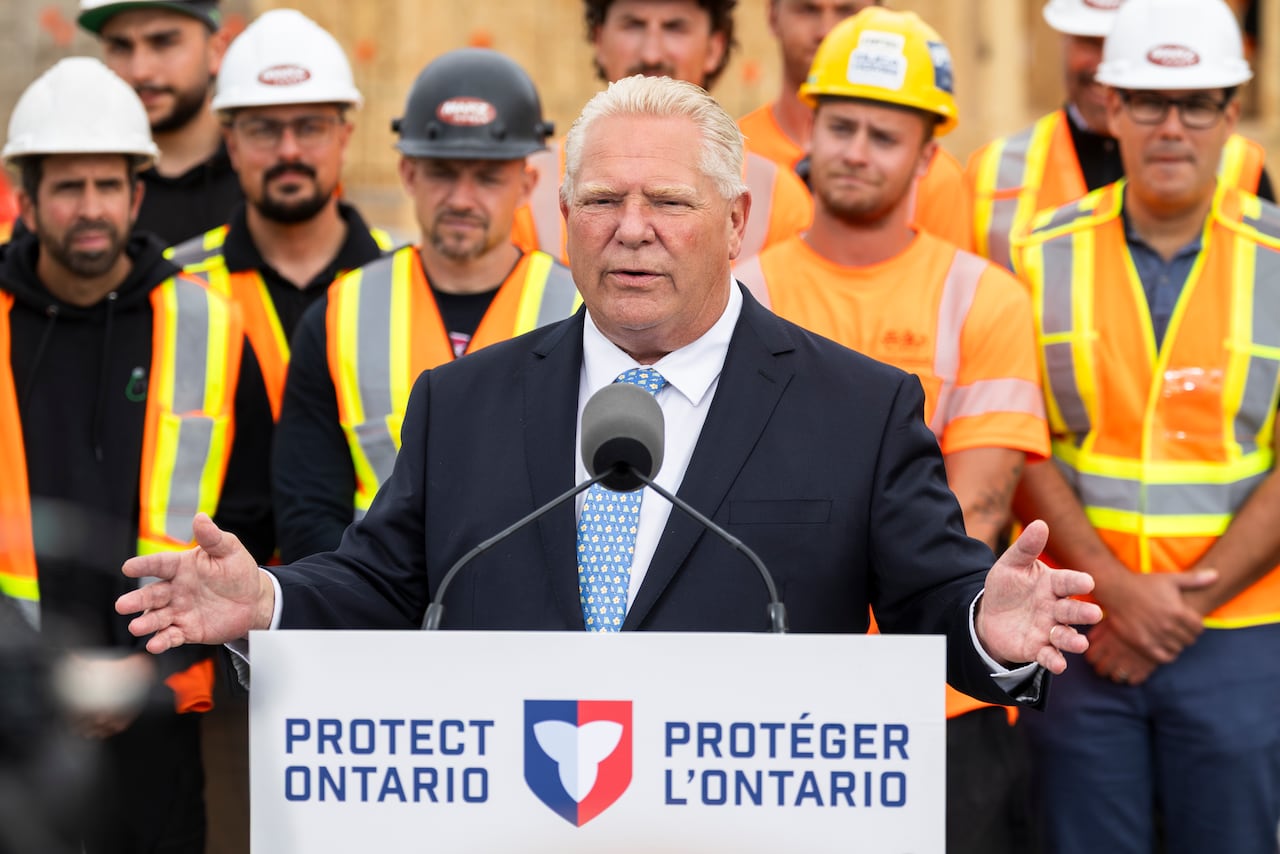 Premier Doug Ford wears a suit and speaks at a podium that says "Protect Ontario" as men wearing hard hats and safety vests stand behind him.