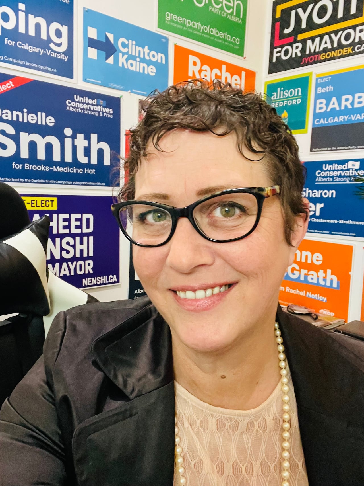 A woman with short hair and glasses sits in a corner adorned with election signs from previous civic, provincial and federal elections in Alberta.