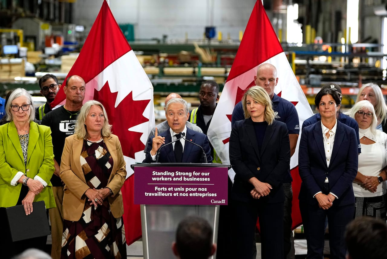A man stands at a podium in front of Canadian flags, flanked by two women on each side