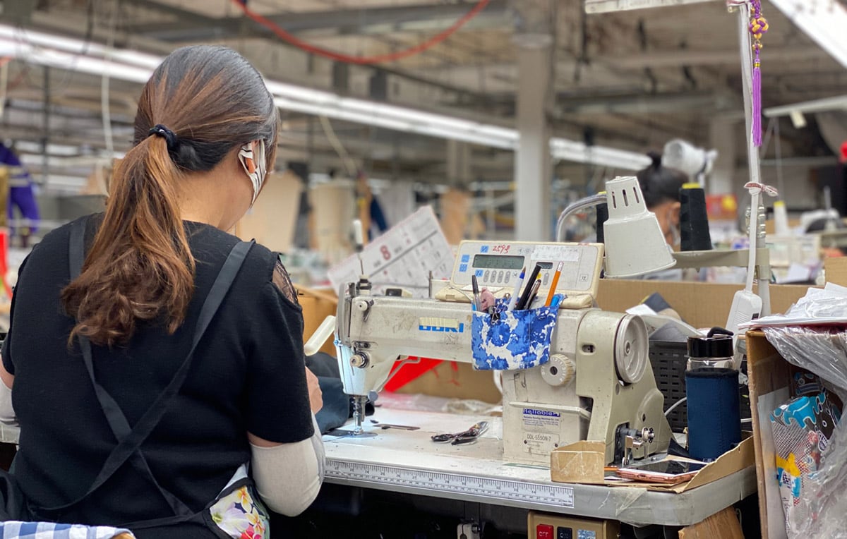 A worker has her back to the camera as she works on a piece of clothing under a sewing machine in a manufacturing warehouse.