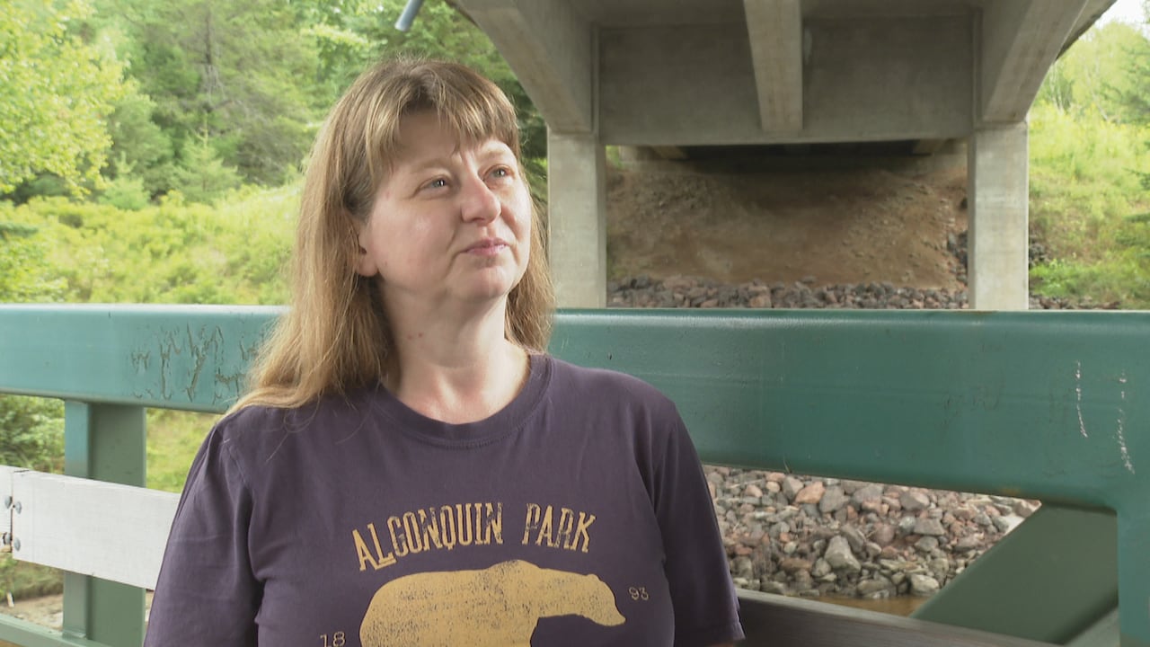 A white blonde woman wearing a navy blue t-shirt with the words 'Algonquin Park' stands under a bridge.