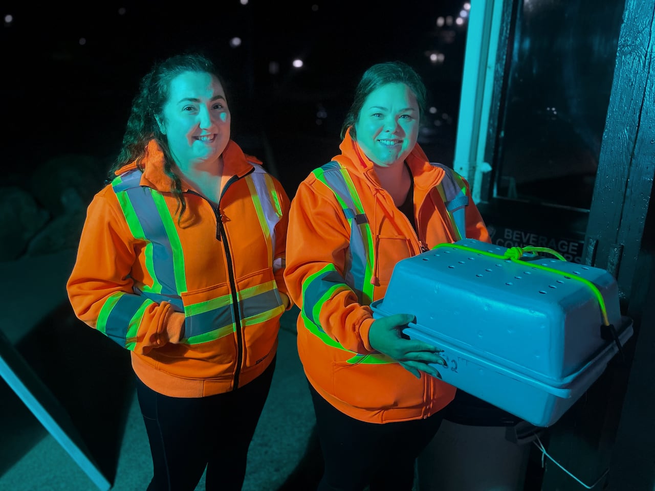 Two women pose in safety vests, one is holding a grey crate with holes on the top.