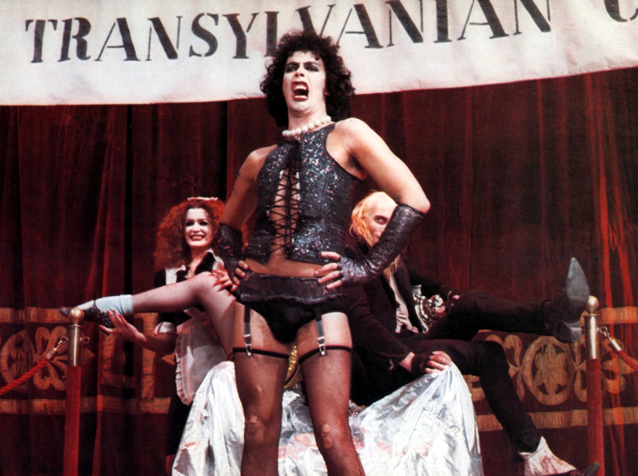 A picture of a cross-dressing man wearing fishnets, black leather gloves, a leather bustier, white pearl necklace and makeup