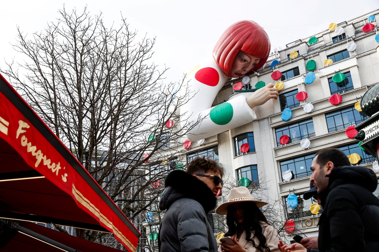 A oversized mannequin of a woman with orange hair is draped over the side of a building. Brightly coloured polka dots are placed on the building.