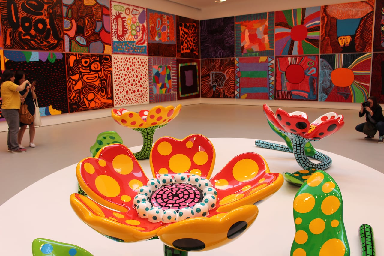 A picture of visitors in a museum looking at paintings on the walls covered in polka dots and swirls, with some colourful flower sculptures in the foreground covered in polkadots.