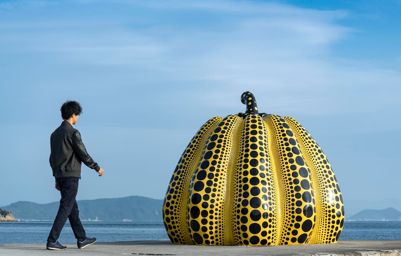 A tourist looking at a yellow pumkin installation beside a body of water
