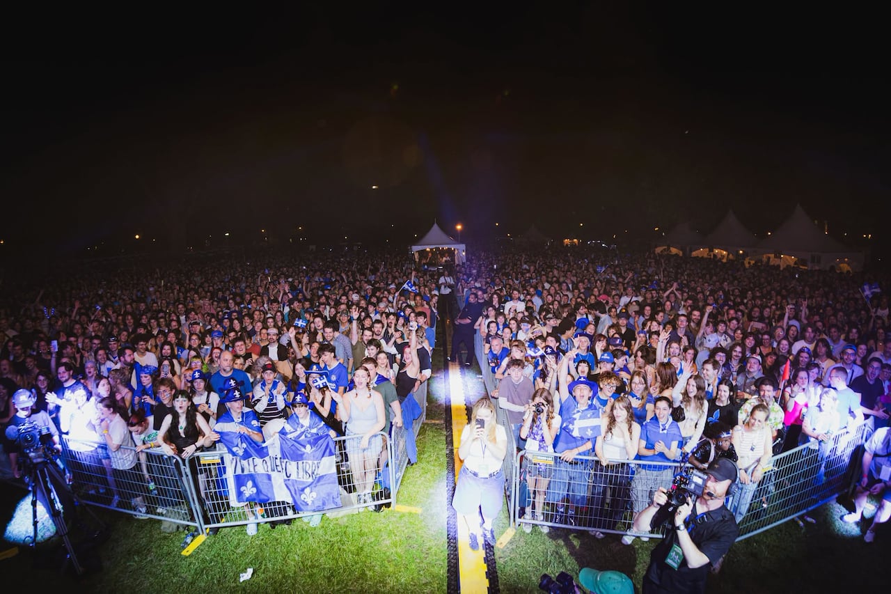 Crowds stand in a field for a concert, with a number of Quebec flags visible