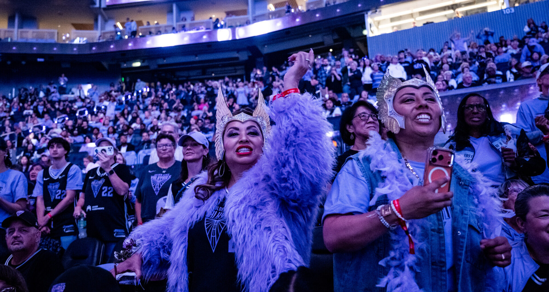 two women in headdresses cheer in stadium with blue and purple light
