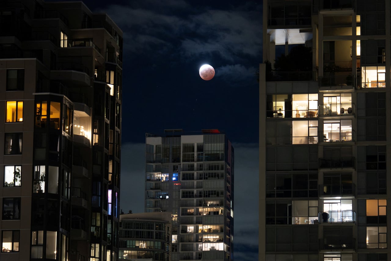 A rising full moon in the sky, seen between buildings at night.