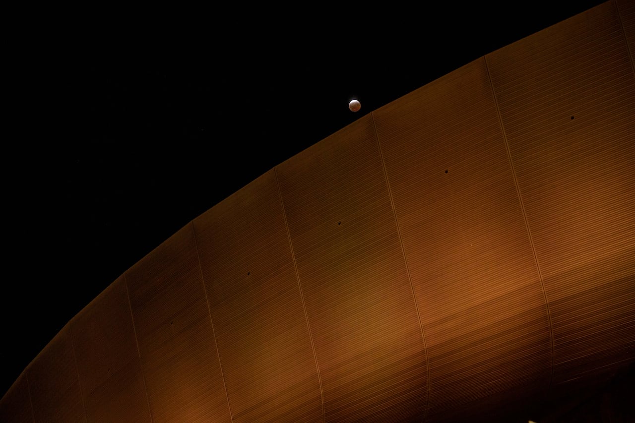 A full moon during a partial lunar eclipse, rising above the curved edge of a stadium at night.