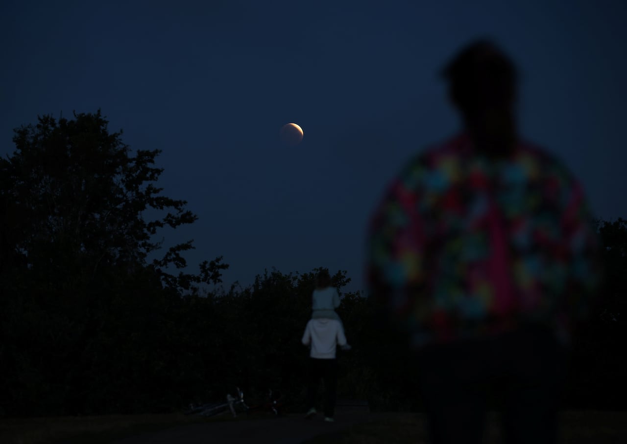The backs of people as they watch a rising full moon in the dark.