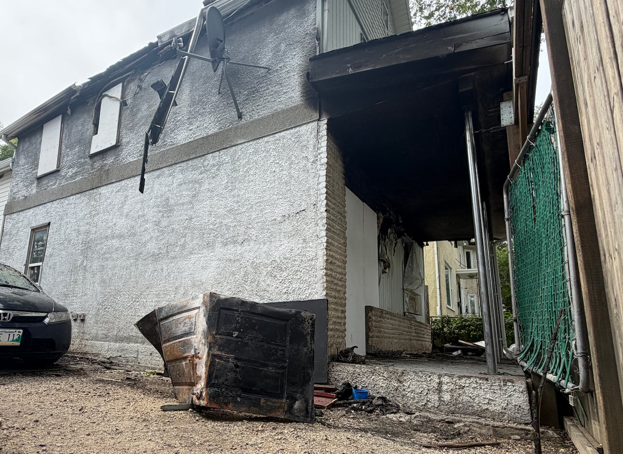 A burned garbage bin sits outside a home with boarded-up windows and signs of smoke damage.